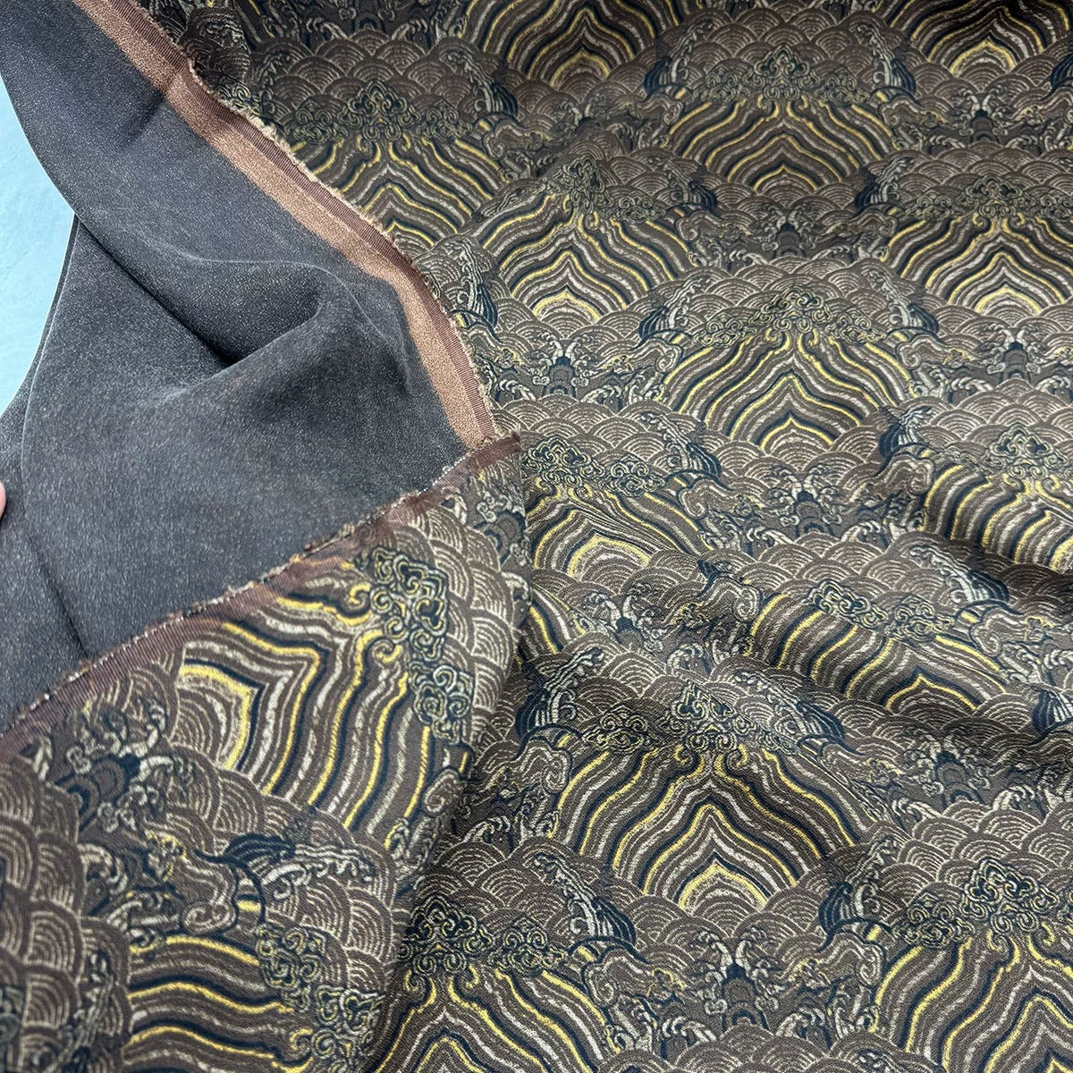 a close-up view of a fabric with a repeating pattern of waves and swirls in shades of brown and gray. The fabric appears to be made of a dark-colored material, possibly a type of textile or upholstery. The pattern is intricate and detailed, with the waves and swirls varying in size and shape, creating a visually striking and textured appearance.