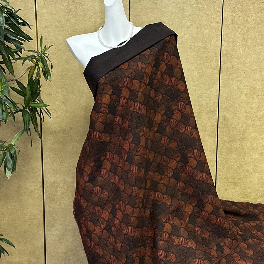 A mannequin wearing a brown and black patterned fabric draped over its body, displayed against a beige wall with a plant in the background.