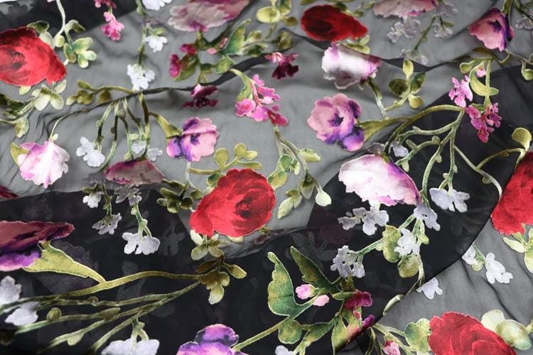 a close-up view of a fabric with a floral pattern. The fabric is predominantly black, with red and white flowers and leaves scattered across it. The flowers appear to be in various stages of bloom, creating a vibrant and lively visual effect. The fabric is described as having a sheer appearance, which adds to the overall delicate and intricate design.