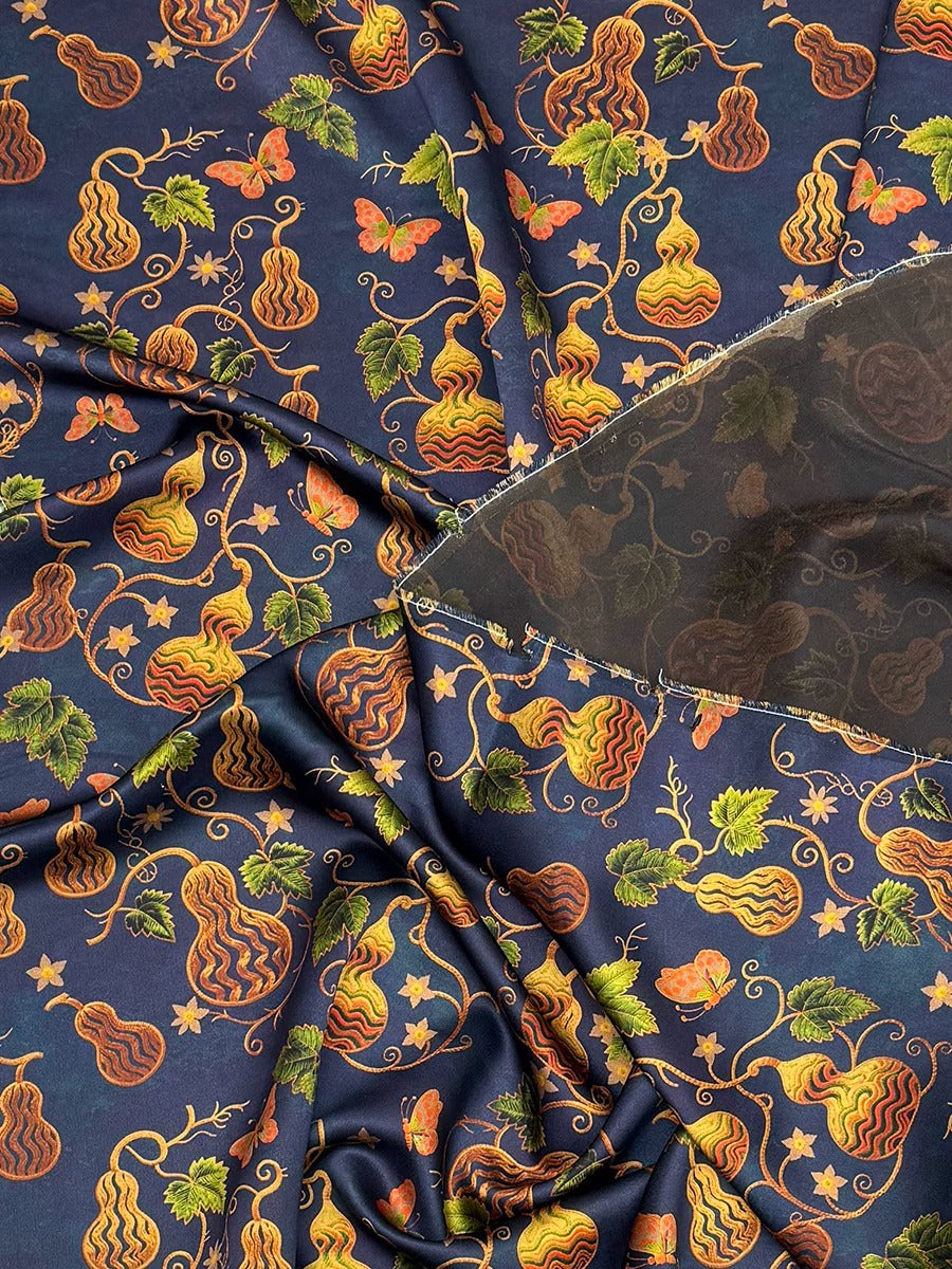 a close-up view of a fabric with a vibrant, colorful pattern of fruits and leaves, including pears, grapes, and leaves in shades of orange, yellow, and green. The fabric appears to be made of a shiny, metallic material, and there is a clear, transparent object partially visible in the bottom right corner of the image.