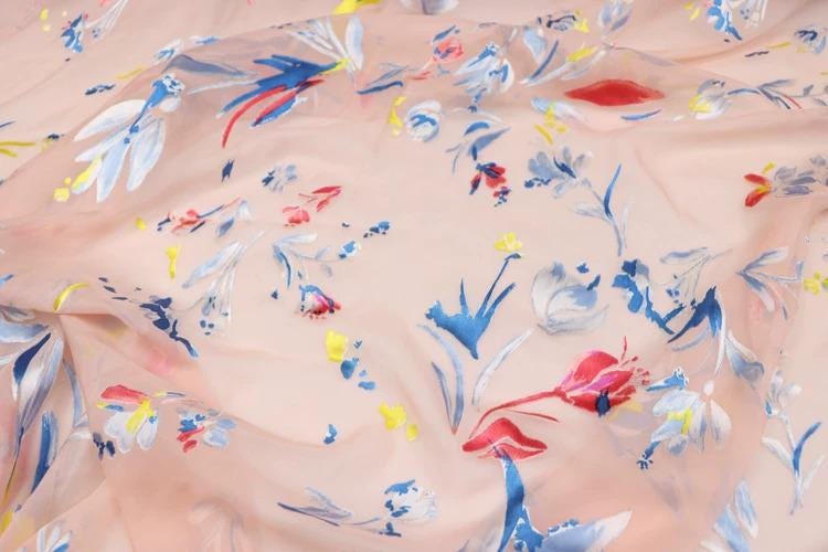a close-up view of a fabric with a floral pattern. The fabric appears to be a light pink color, and the pattern consists of various flowers and leaves in different colors, including red, blue, and yellow. The fabric is crumpled, giving it a soft and delicate appearance.