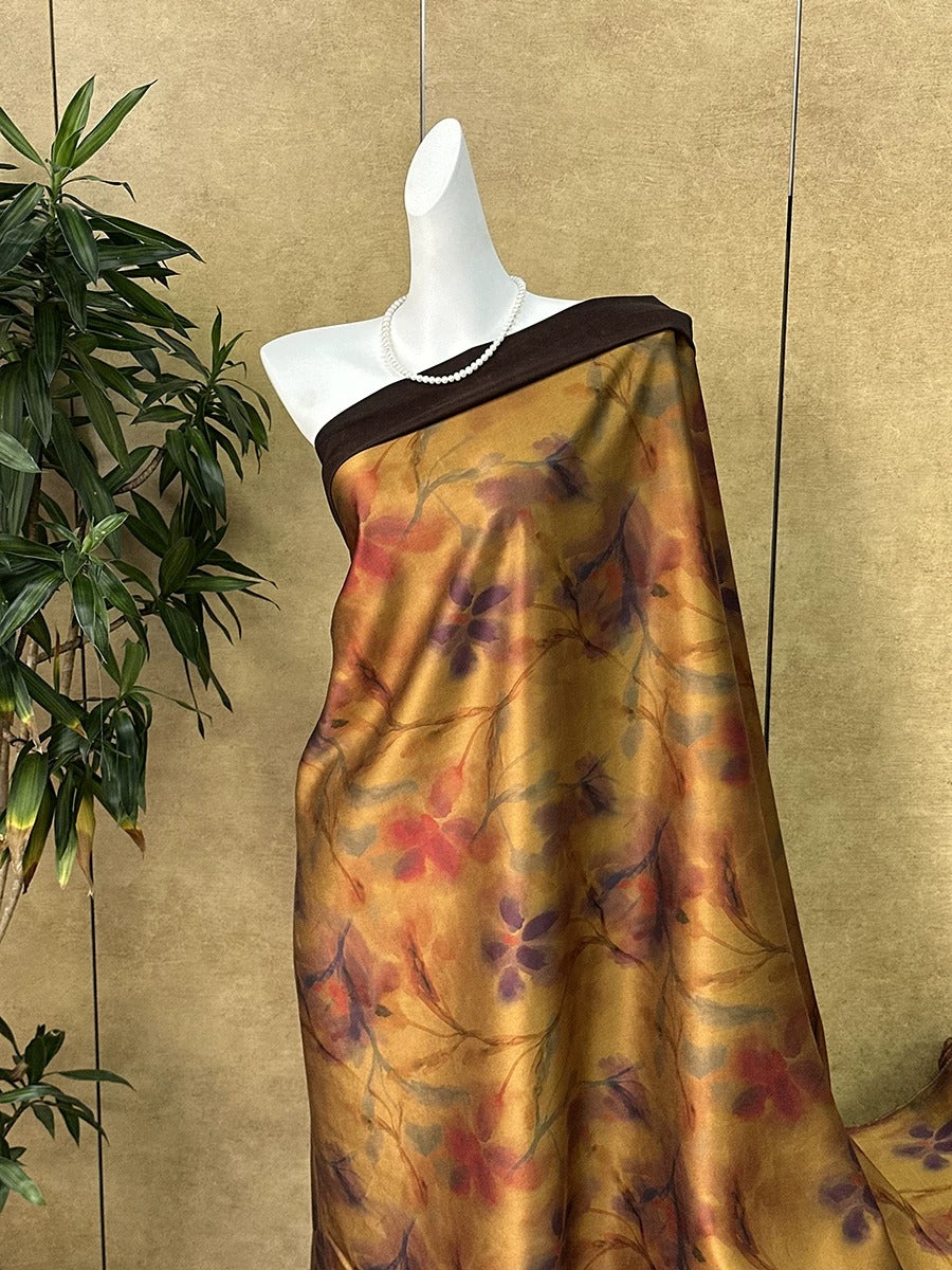 A mannequin wearing a gold and purple floral print dress with a brown waistband, displayed against a beige wall with a plant in the background.
