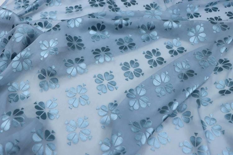 a close-up view of a fabric with a repeating pattern of blue and white flowers or leaves. The pattern is intricate and detailed, with each flower or leaf being a small, symmetrical design. The fabric appears to be made of a soft, flowing material, such as chiffon or silk, and has a slightly wrinkled texture. The pattern repeats itself across the entire fabric, creating a sense of depth and dimension.