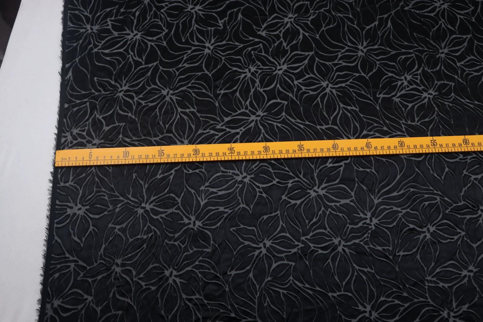 a close-up view of a fabric with a black and gray floral pattern. A yellow measuring tape is laid across the fabric, indicating the size or dimensions of the fabric.