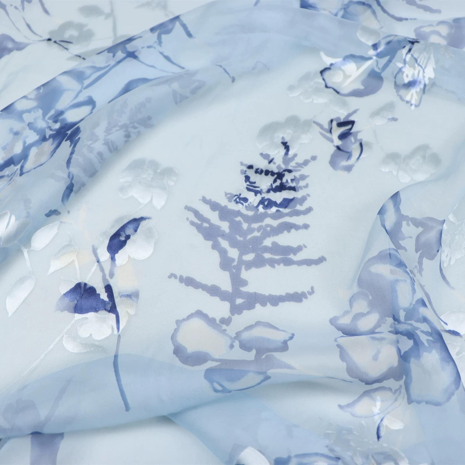 The image is a close-up of a fabric with a blue floral pattern. The fabric appears to be made of sheer material, with a delicate and intricate design featuring various shades of blue and white flowers and leaves. The fabric is crumpled, giving it a soft and delicate appearance.