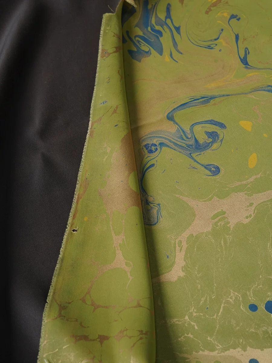 a close-up view of a fabric with a marbled pattern in shades of green and blue. The fabric appears to be a piece of clothing or textile, with a dark background.