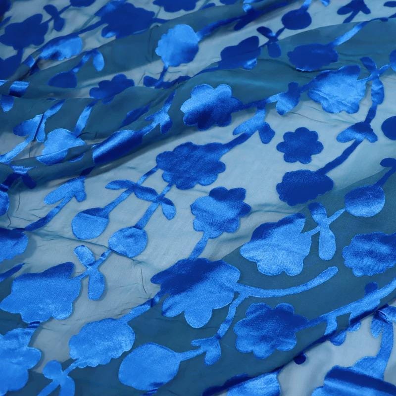 a close-up view of a fabric with a blue floral pattern. The fabric appears to be made of a shiny, silky material and has a smooth texture. The pattern consists of blue flowers and leaves, which are the main decorative elements on the fabric. The fabric is spread out, revealing the intricate design and the vibrant blue color of the flowers and leaves.