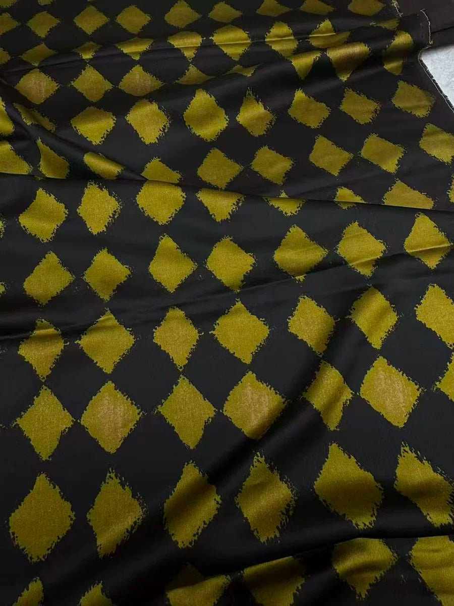 a close-up view of a fabric with a black and gold diamond pattern. The diamonds are arranged in a diagonal pattern, creating a visually striking design. The fabric appears to be made of a shiny material, possibly silk or satin, which adds to the overall aesthetic of the image.