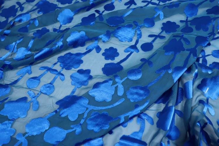 a close-up view of a fabric with a blue floral pattern. The fabric appears to be made of a shiny, silky material and has a textured, wrinkled appearance. The pattern consists of blue flowers and leaves, creating a visually striking and intricate design.