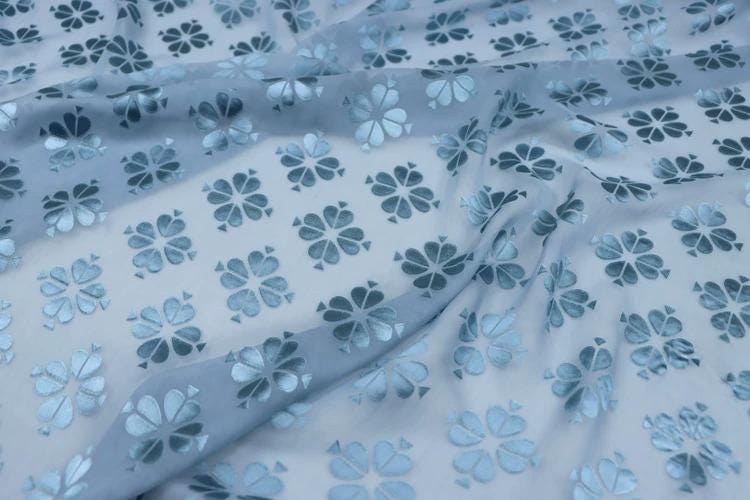 a close-up view of a fabric with a repeating pattern of blue and white flowers or leaves. The fabric appears to be made of a soft, flowing material, and the pattern is quite intricate and detailed.