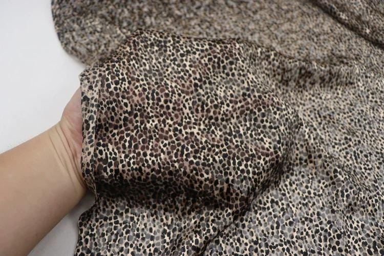A hand is holding a fabric with a leopard print pattern, which appears to be a close-up view of the fabric.