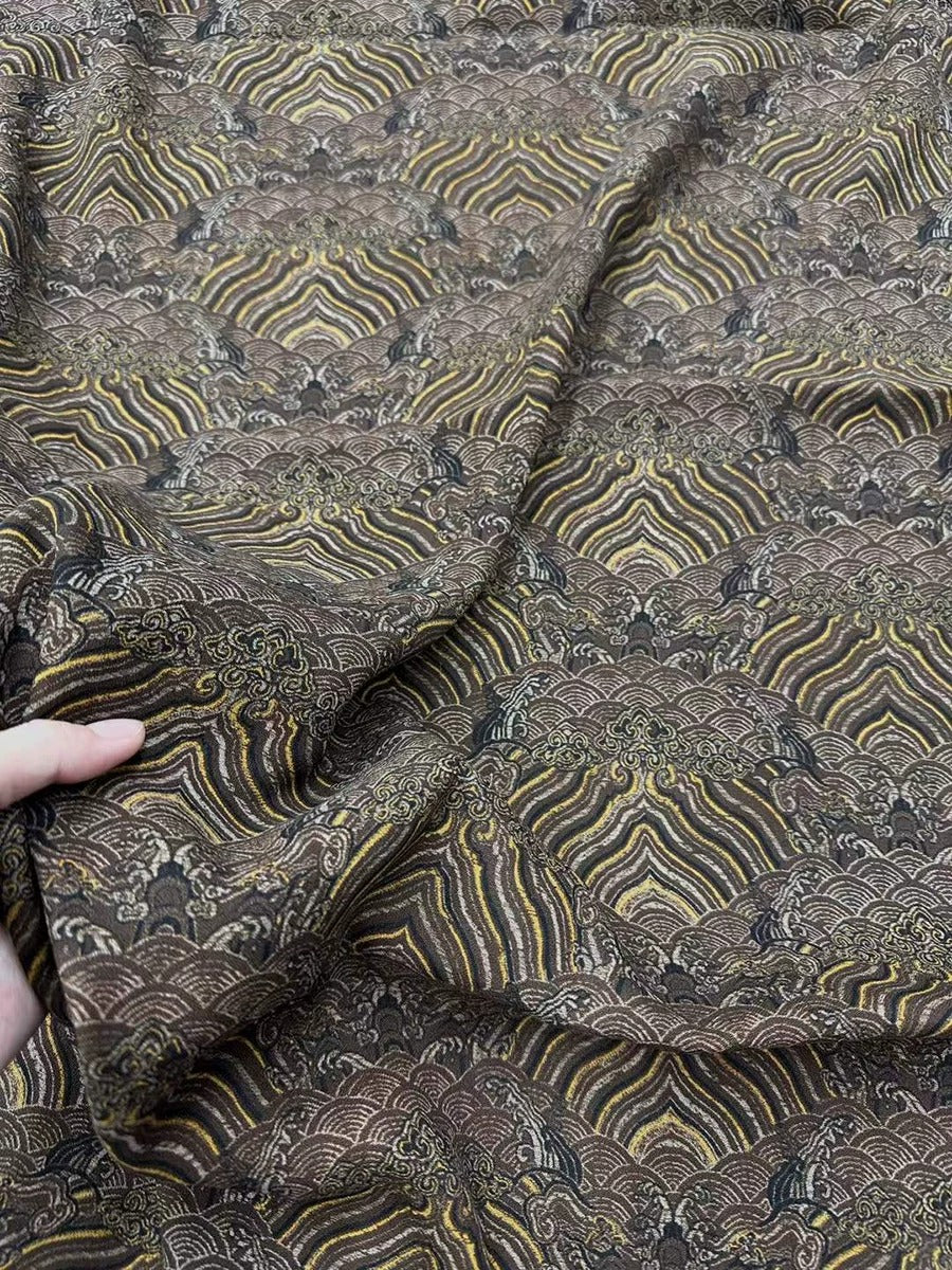 a close-up view of a fabric with a repeating pattern of waves and swirls in shades of brown and gold. The fabric appears to be made of a soft, textured material, and the intricate design suggests it could be a piece of clothing or textile.