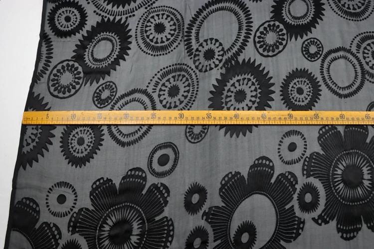 a close-up view of a fabric with a black and white floral pattern. A yellow measuring tape is placed across the fabric, indicating the size of the fabric.