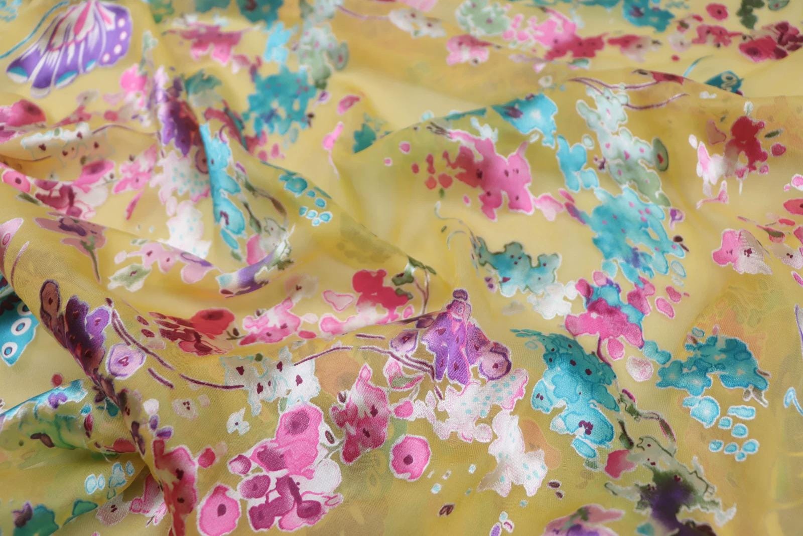 The image is a close-up of a fabric with a vibrant, colorful floral pattern. The fabric appears to be made of silk or a similar material, and the pattern consists of various shades of yellow, pink, blue, and green flowers and leaves. The fabric is crumpled, giving it a soft, textured appearance.