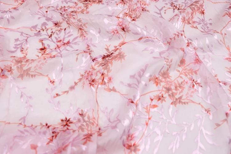 a close-up view of a fabric with a floral pattern in shades of pink and white. The fabric appears to be made of sheer material, giving it a delicate and airy appearance. The pattern consists of various pink and white flowers and leaves, creating a visually appealing and intricate design.