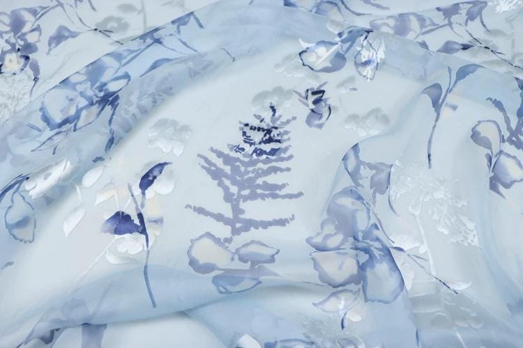 a close-up view of a fabric with a blue floral pattern. The fabric appears to be made of sheer material, with a delicate and intricate design. The pattern consists of various shades of blue and white, creating a visually appealing and harmonious composition.