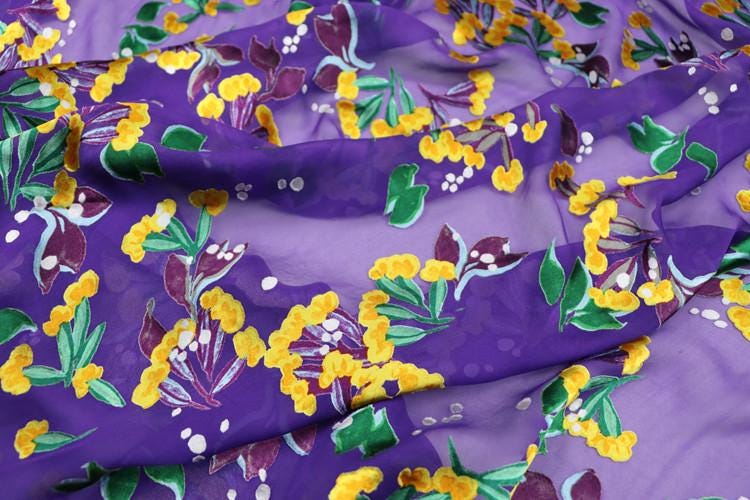 a close-up view of a fabric with a vibrant floral pattern in shades of purple and yellow. The pattern features various flowers and leaves, creating a visually striking and colorful design.