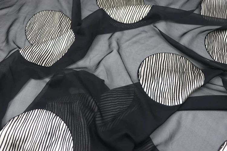 a close-up view of a fabric with a black and white striped pattern. The fabric appears to be made of a sheer material, giving it a soft and flowing appearance. The pattern consists of circular shapes with diagonal stripes, creating a visually striking and intricate design.