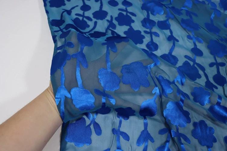 a close-up view of a fabric with a blue floral pattern. The fabric appears to be made of a sheer material, and the pattern consists of blue flowers and leaves. The fabric is folded and held in the hand, allowing for a detailed view of the intricate design.