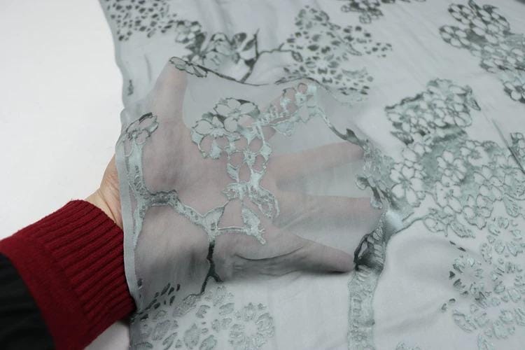 A hand is holding a piece of fabric with a floral pattern, which appears to be a light gray color.