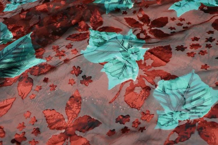 a close-up view of a fabric with a floral pattern. The fabric has a mix of red and green colors, with various leaf and flower designs scattered across the surface. The pattern appears to be a combination of leaves and flowers, creating a visually appealing and intricate design.