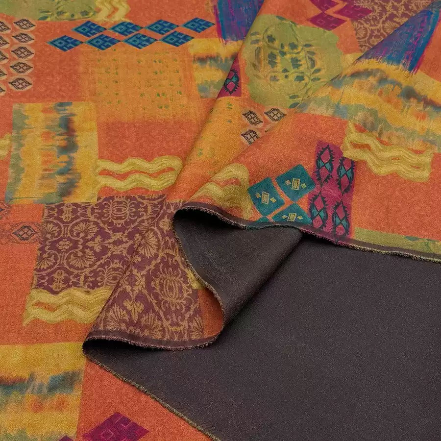 a vibrant, colorful fabric with a pattern of geometric shapes and designs in shades of orange, yellow, and purple. The fabric appears to be a textile, possibly a blanket or a garment, with a dark-colored underside.