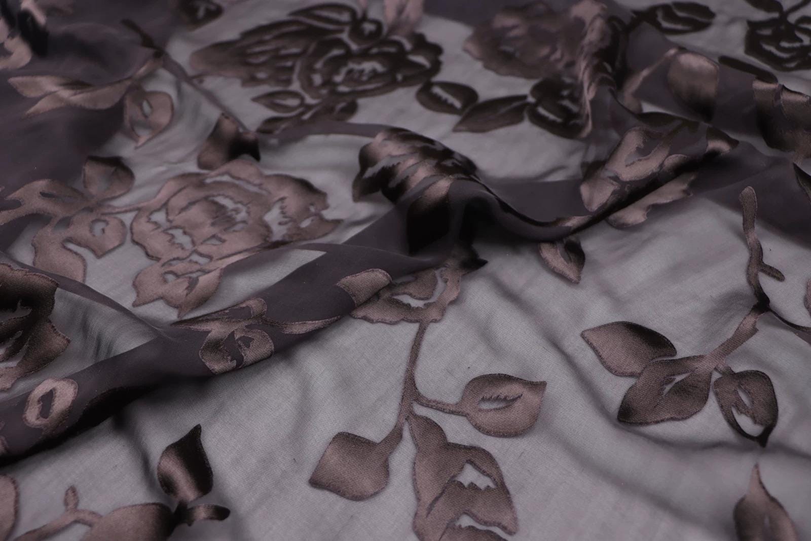 a close-up view of a fabric with a dark, intricate floral pattern. The fabric appears to be made of a shiny, dark material, possibly silk or satin, and features a repeating pattern of leaves and flowers. The image does not contain any text or other discernible objects, and the focus is solely on the detailed floral design on the fabric.