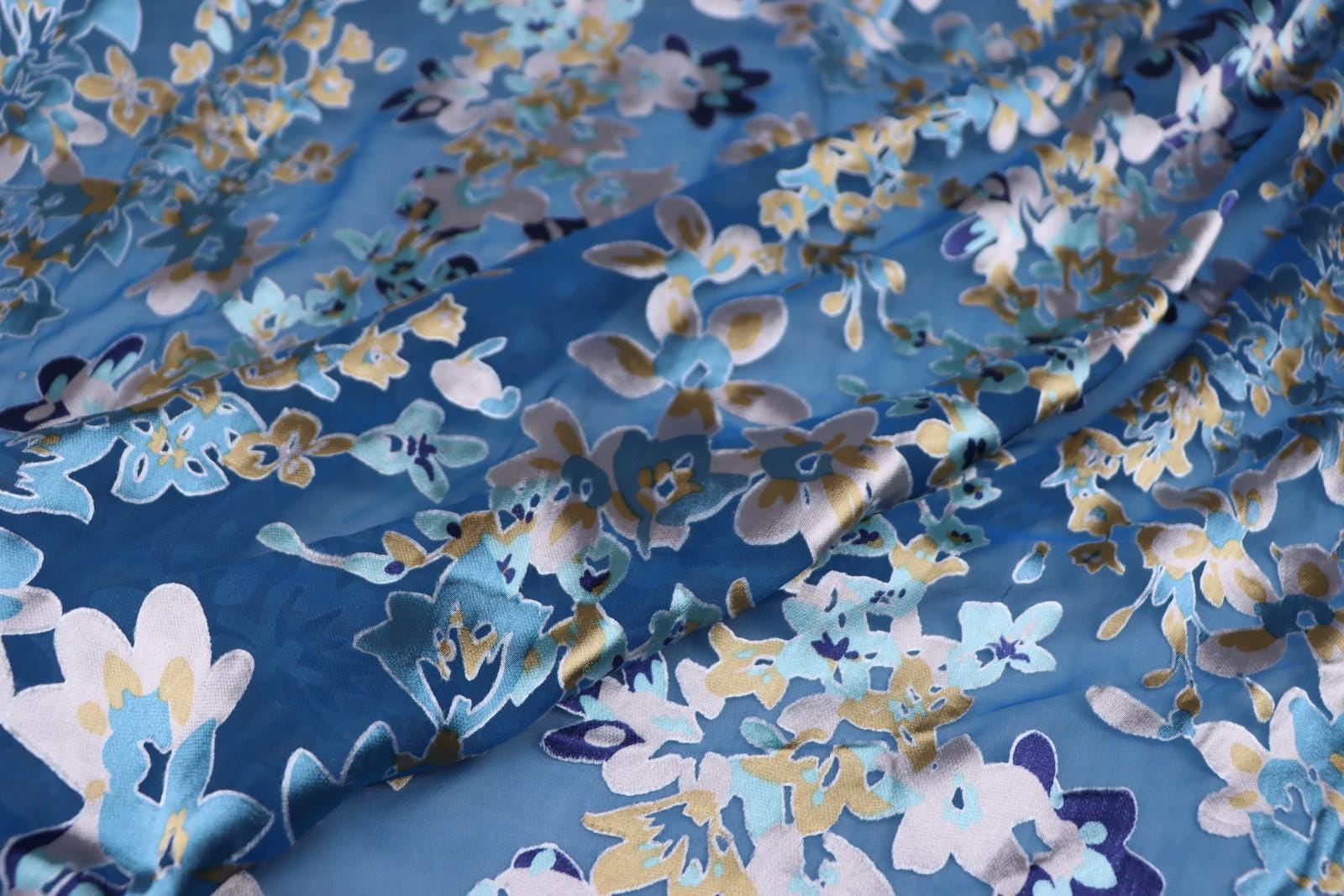 a close-up view of a fabric with a floral pattern in shades of blue, white, and gold. The fabric appears to be made of silk or a similar material, and the pattern is intricate and detailed.