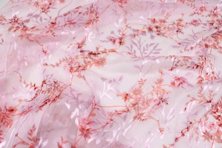 a close-up view of a fabric with a pink floral pattern. The fabric appears to be made of sheer material, with delicate pink flowers and leaves covering the entire surface. The pattern is intricate and detailed, creating a visually appealing and textured appearance.