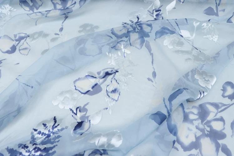 a close-up view of a fabric with a blue floral pattern. The fabric appears to be a sheer material, with a light blue background and white flowers and leaves printed on it. The pattern is intricate and detailed, showcasing the craftsmanship of the fabric.