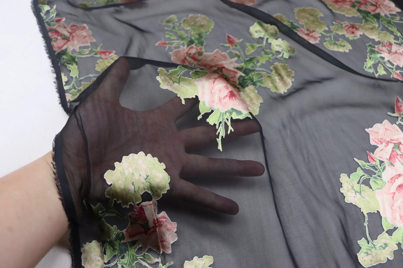 A hand is holding a piece of fabric with a floral pattern, which appears to be a black sheer material.