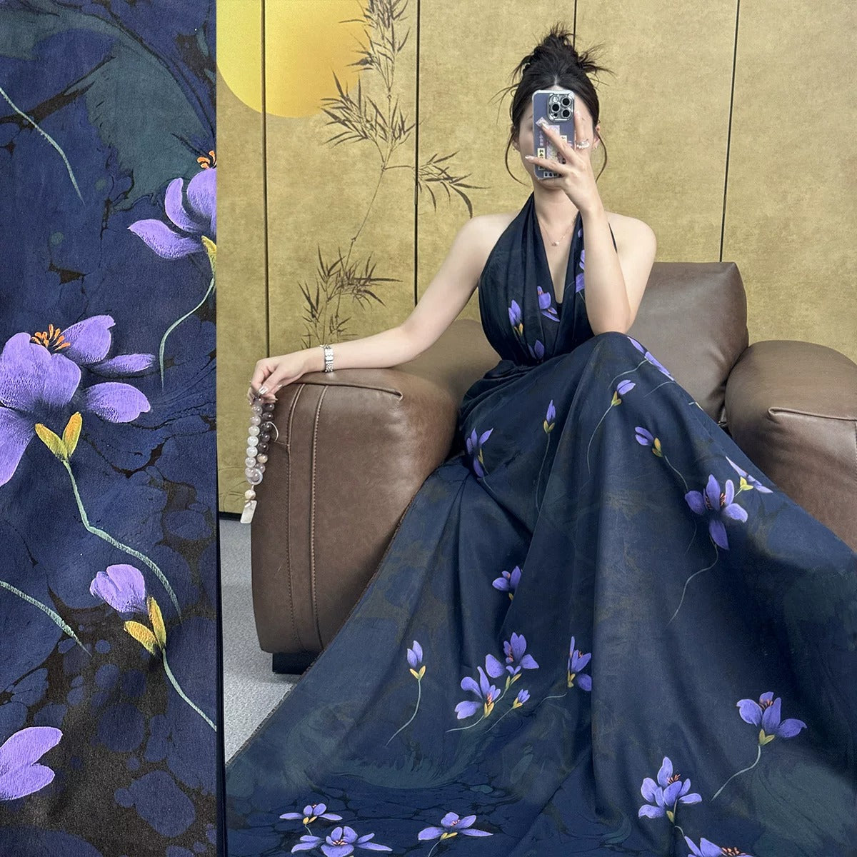 A woman in a long, dark blue dress with purple flowers is sitting on a brown leather couch, holding a camera and taking a selfie.