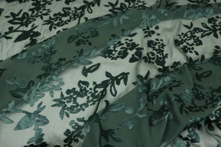 a close-up view of a fabric with a floral pattern in shades of green and gray. The fabric appears to be made of silk or a similar material, and the pattern consists of various leaf and flower designs. The fabric is crumpled, giving it a textured appearance.
