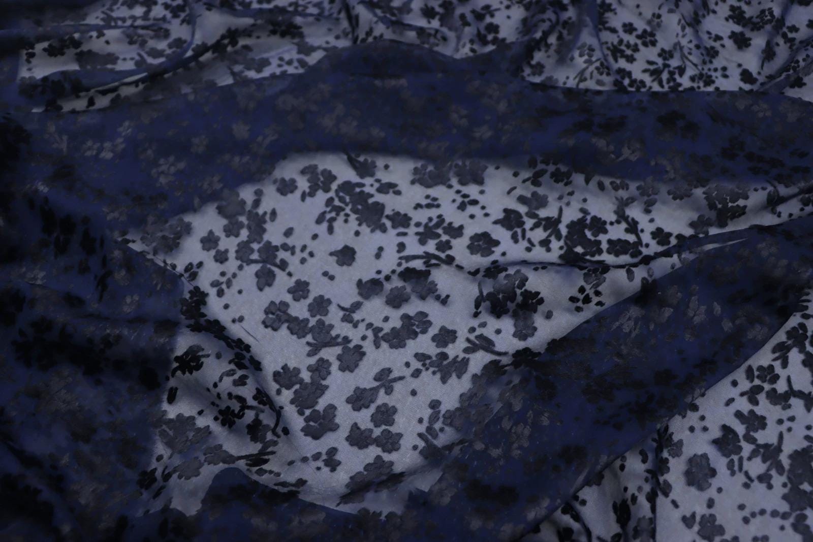 a close-up view of a fabric with a floral pattern in shades of blue and white. The fabric appears to be made of sheer material, with a dark blue background and a lighter blue floral pattern.