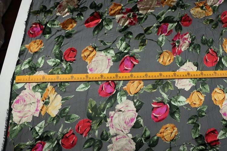 a fabric with a floral pattern, featuring a variety of colorful flowers such as roses, lilies, and other blooms. The fabric appears to be gray, and there is a yellow measuring tape laid out on it, indicating that the fabric is being measured for a specific purpose.