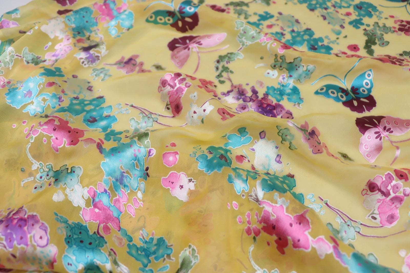 a close-up view of a fabric with a vibrant, colorful floral pattern. The fabric appears to be made of silk or a similar material, and it features a variety of flowers and butterflies in shades of pink, blue, green, and yellow. The pattern is intricate and detailed, with each flower and butterfly adding to the overall visual appeal of the fabric.
