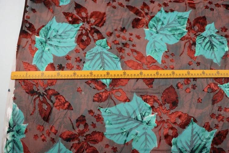 a fabric with a floral pattern of red and green leaves, and a yellow measuring tape is placed across the fabric to measure its length.