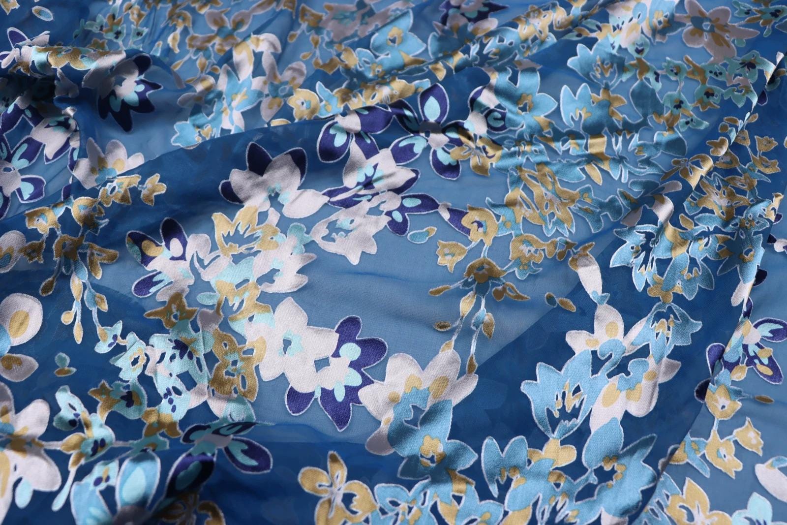 a close-up view of a fabric with a blue and white floral pattern. The fabric appears to be made of silk or a similar material, and the pattern consists of various shades of blue and white flowers and leaves. The fabric is crumpled, giving it a textured appearance.