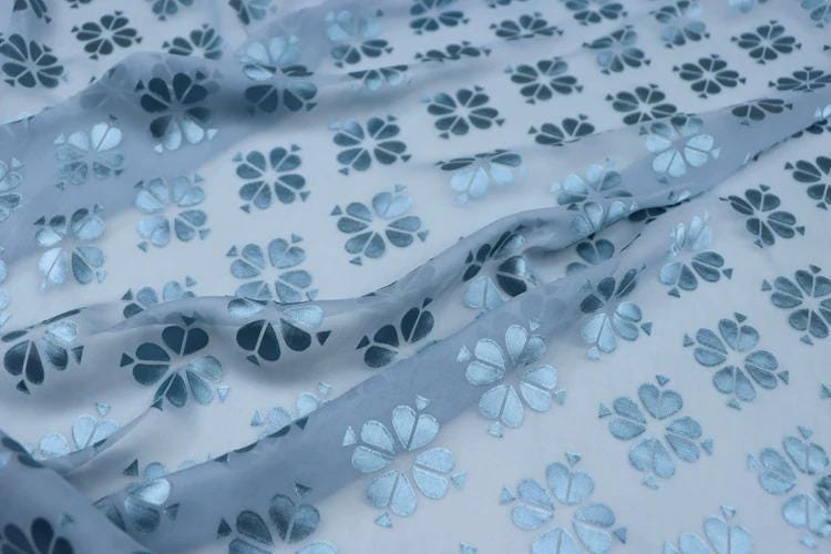 a close-up view of a fabric with a repeating pattern of blue and white flowers. The fabric appears to be made of a soft, flowing material, and the pattern is intricate and detailed.