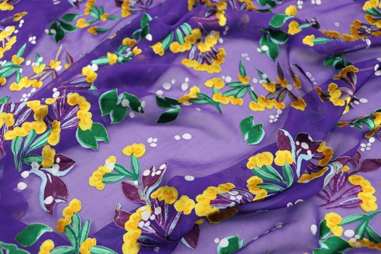 a close-up view of a fabric with a vibrant floral pattern in shades of purple, yellow, and green. The pattern features various flowers and leaves, creating a lively and colorful design.