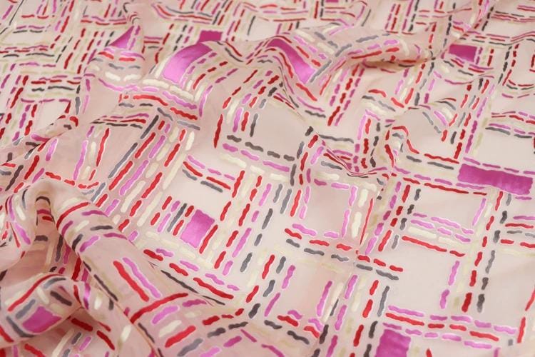 a close-up view of a fabric with a geometric pattern of pink, red, and gray lines and shapes. The fabric appears to be made of a soft, light-colored material, possibly silk or chiffon. The pattern is intricate and detailed, with the lines and shapes creating a visually striking design.