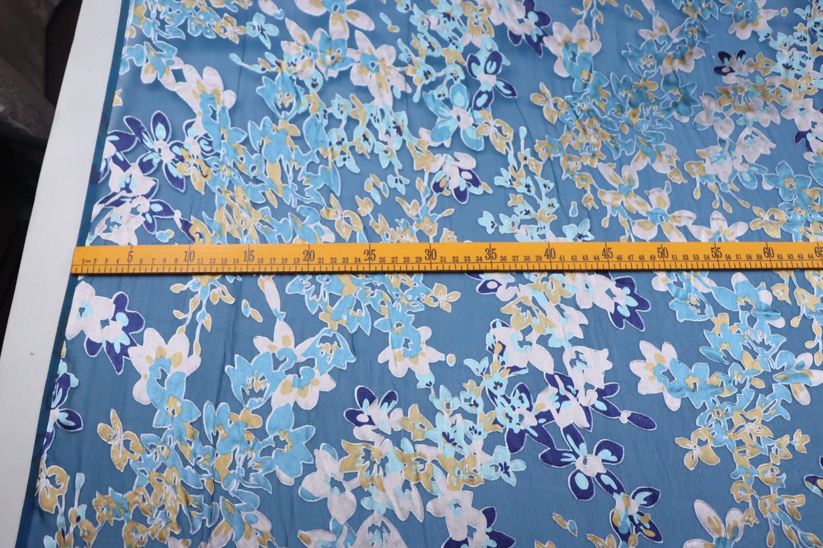 a fabric with a blue and white floral pattern, and a yellow measuring tape is placed across it, indicating the size or dimensions of the fabric.