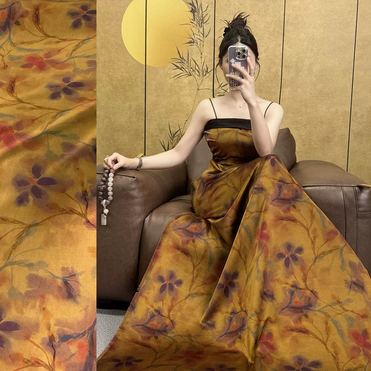 A woman in a long, floral-patterned dress is sitting on a leather couch, holding a necklace and taking a selfie with her phone.