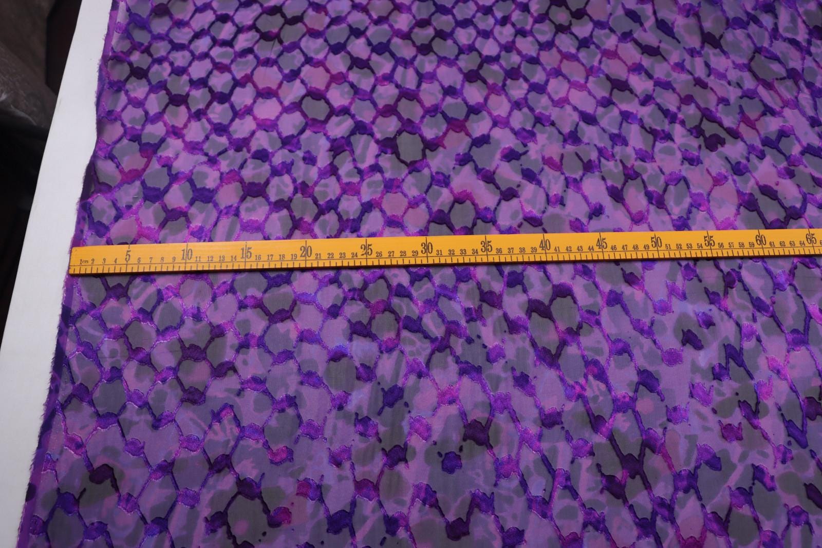 a fabric with a purple and gray pattern, and a yellow measuring tape is placed across it, indicating the size of the fabric.
