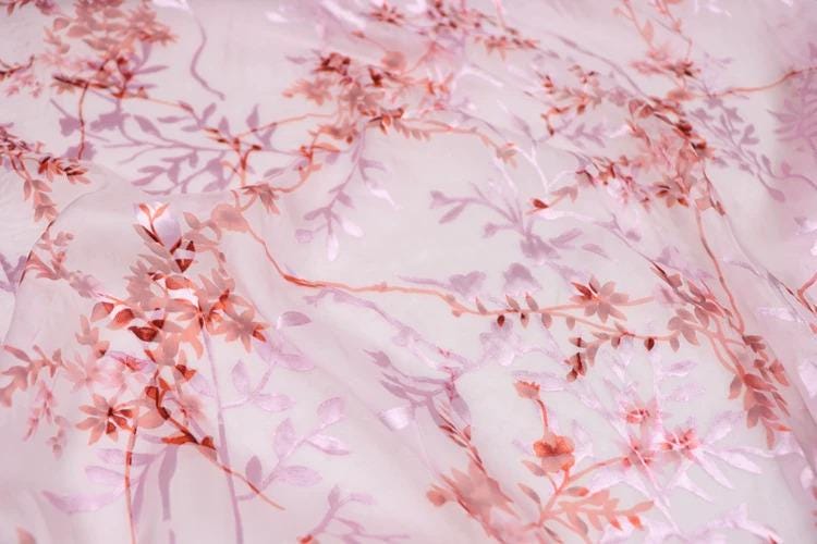 a close-up view of a fabric with a floral pattern in shades of pink and red. The fabric appears to be made of sheer material, giving it a delicate and airy appearance. The pattern consists of various floral elements, such as leaves and flowers, arranged in a visually appealing manner. The image does not contain any text or other discernible objects, and the focus is solely on the intricate floral design on the fabric.