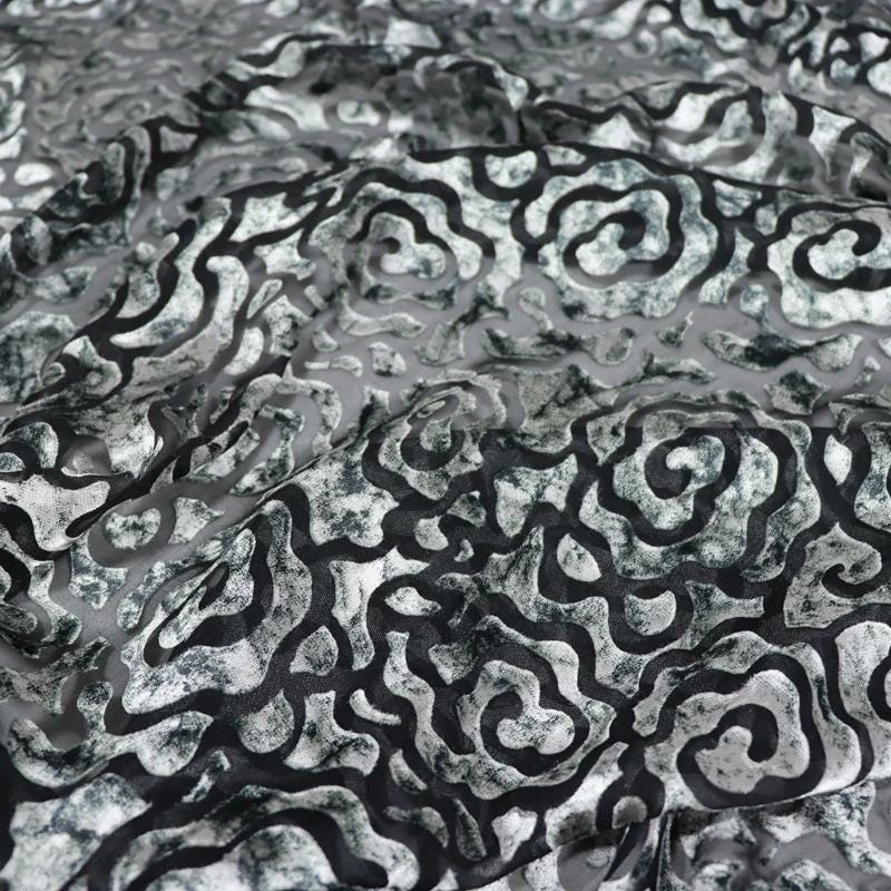 a close-up view of a fabric with a repeating pattern of black and white swirls or curves. The pattern is intricate and detailed, creating a visually striking and textured appearance.