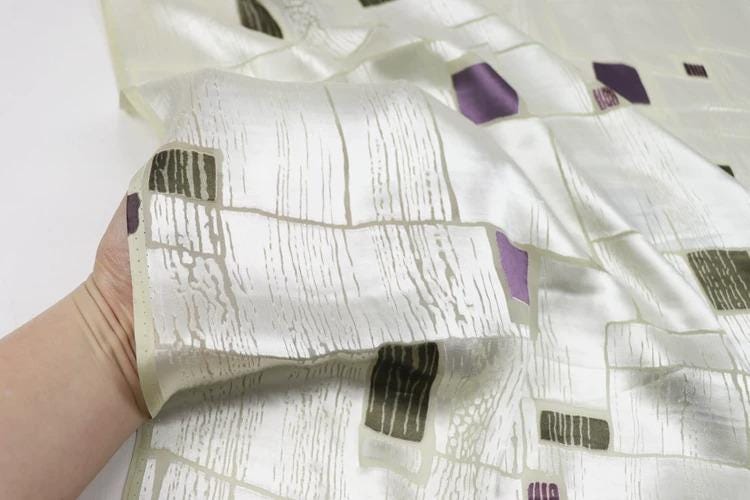 A hand is holding a piece of fabric with a pattern of white and purple squares and rectangles.