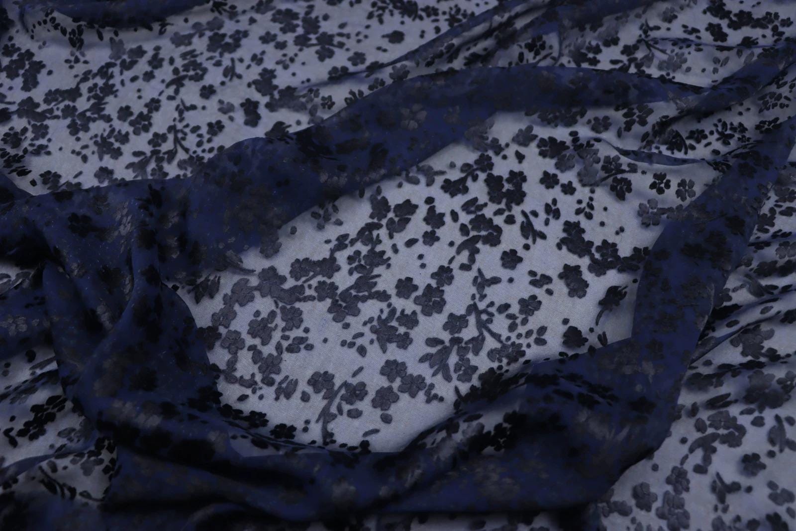 a close-up view of a fabric with a floral pattern in shades of blue and white. The fabric appears to be made of sheer material, with a dark blue color dominating the background. The pattern consists of small black flowers or leaves scattered across the fabric, creating a visually striking contrast against the lighter background.
