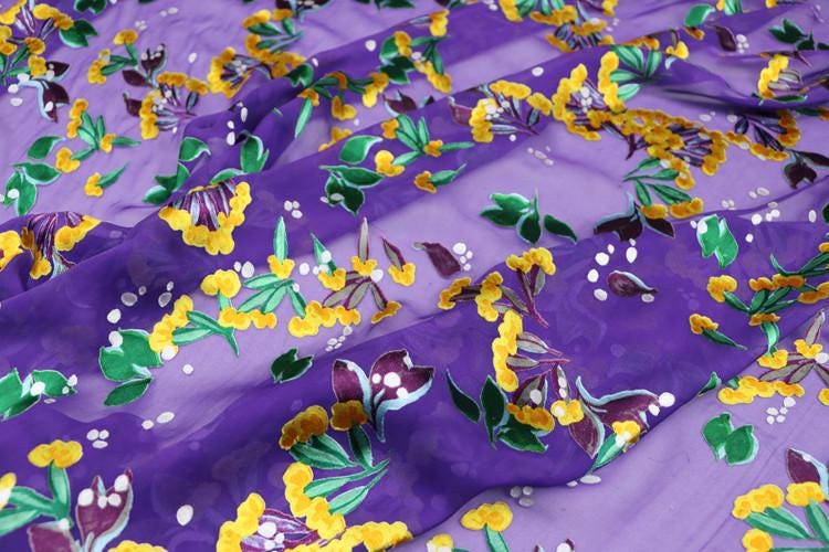 a close-up view of a fabric with a vibrant floral pattern in shades of purple and yellow. The pattern features various flowers and leaves, creating a lively and colorful design.