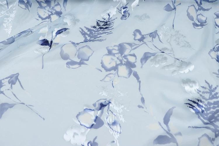 a close-up view of a fabric with a floral pattern in shades of blue and white. The pattern consists of various flower-like shapes and leaves, creating a visually appealing and intricate design.